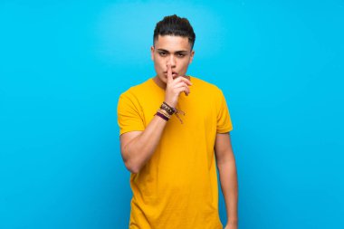Young man with yellow shirt over isolated blue background showing a sign of silence gesture putting finger in mouth