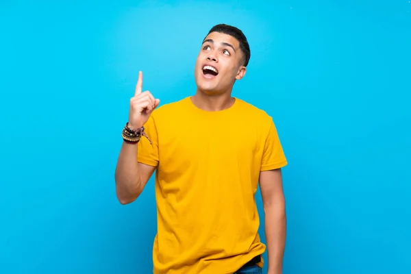 Young man with yellow shirt over isolated blue background pointing up and surprised
