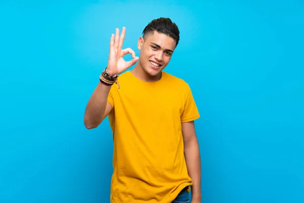 Young man with yellow shirt over isolated blue background showing ok sign with fingers