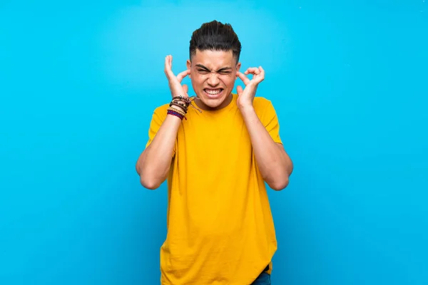 Young man with yellow shirt over isolated blue background frustrated and covering ears