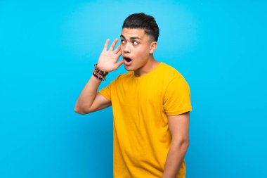 Izole mavi arka plan üzerinde sarı gömlekli genç adam kulağına el koyarak bir şey dinlerken