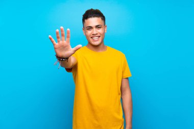 Young man with yellow shirt over isolated blue background counting five with fingers