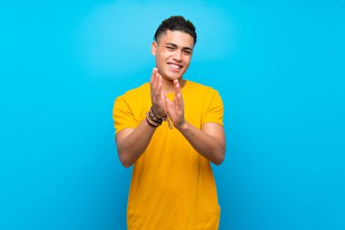 Young man with yellow shirt over isolated blue background applauding after presentation in a conference