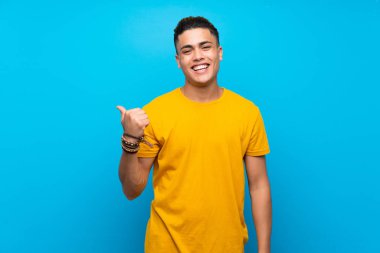 Izole mavi arka plan üzerinde sarı gömlek li genç adam bir ürün sunmak için yan işaret