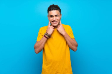 Young man with yellow shirt over isolated blue background smiling with a happy and pleasant expression