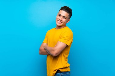 Young man with yellow shirt over isolated blue background with arms crossed and looking forward
