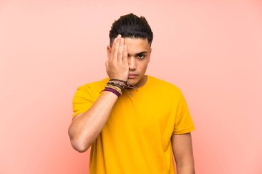 Young man with yellow shirt over isolated pink wall covering a eye by hand