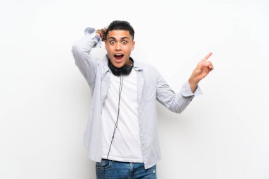 Young man over isolated white wall with earphones surprised and pointing finger to the side