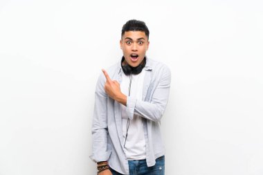 Young man over isolated white wall with earphones surprised and pointing side