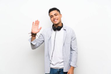 Young man over isolated white wall with earphones saluting with hand with happy expression