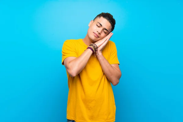 Young man with yellow shirt over isolated blue background making sleep gesture in dorable expression