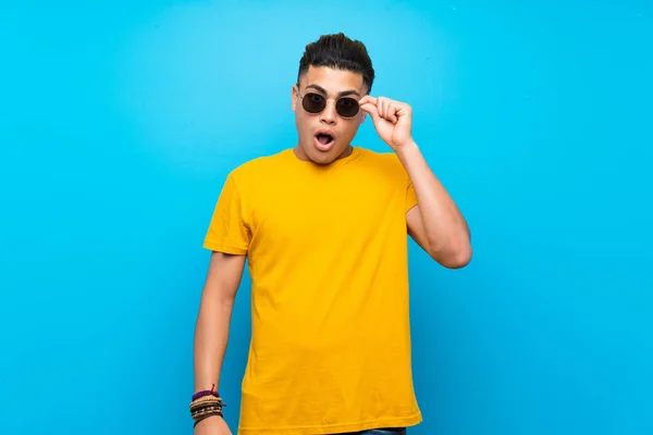 Young man with yellow shirt over isolated blue background with glasses and surprised