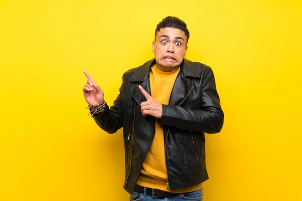 Young man over isolated yellow background frightened and pointing to the side