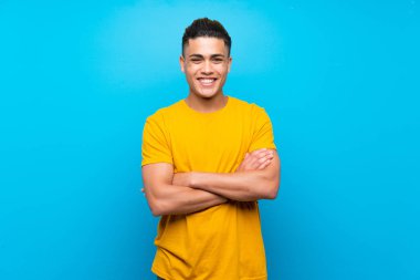 Young man with yellow shirt over isolated blue background keeping the arms crossed in frontal position