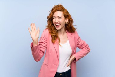 Izole mavi duvar üzerinde takım elbiseli Redhead kadın mutlu ifade ile el ile selamlama