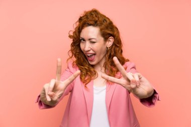 Izole pembe duvar üzerinde takım elbiseredhead kadın gülümseyerek ve zafer işareti gösteren