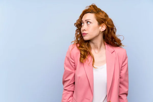 Redhead woman in suit over isolated blue wall making doubts gesture looking side