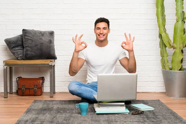 Handsome man sitting on the floor with his laptop showing an ok sign with fingers