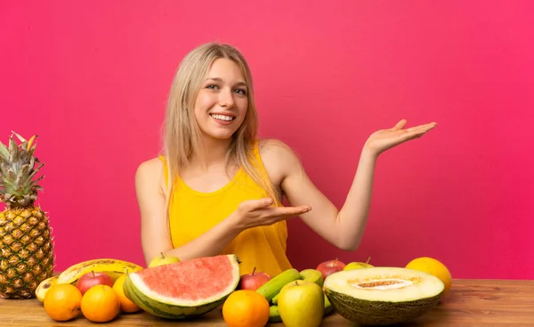 Young blonde woman with lots of fruits extending hands to the side for inviting to come