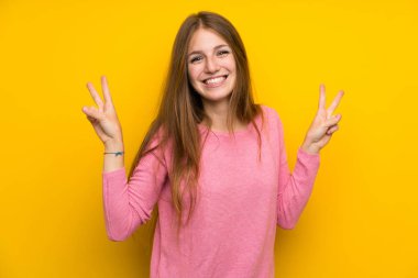 İzole edilmiş sarı duvarın üzerinde uzun saçlı genç bir kadın zafer işaretini iki eliyle gösteriyor.