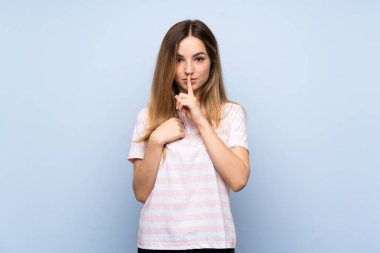 İzole edilmiş mavi arka planda genç bir kadın sessizlik işareti gösteriyor. Parmağını ağzına sok.