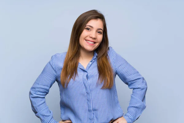 Izole mavi duvar üzerinde genç kız kalça kolları ile poz ve gülümseyerek