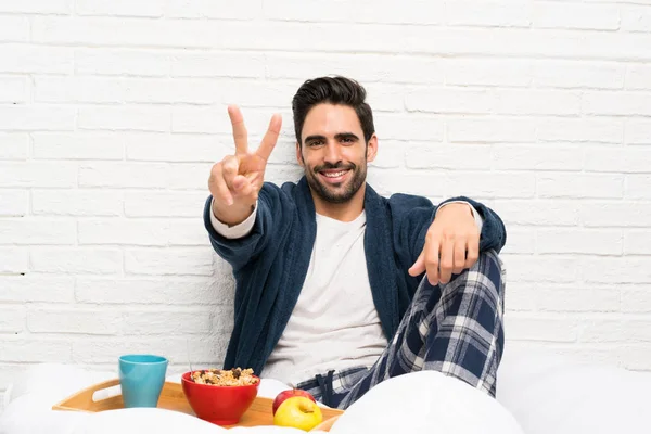 Man in bed with dressing gown and having breakfast smiling and showing victory sign