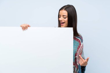 Bir kavram eklemek için boş beyaz bir pankart tutan izole mavi duvar üzerinde genç kadın