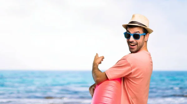 Man with hat and sunglasses on his summer vacation pointing back with the index finger presenting a product from behind at the beach