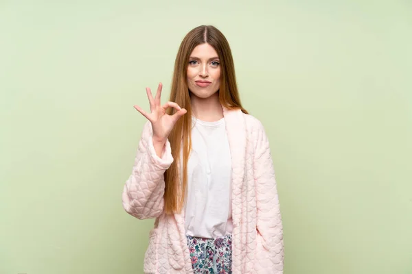 Young woman in dressing gown over green wall showing an ok sign with fingers