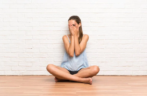 Young woman sitting on the floor covering eyes and looking through fingers
