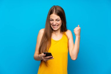 Zafer pozisyonunda telefon ile izole mavi duvar üzerinde uzun saçlı genç kadın