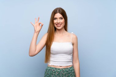 İzole edilmiş mavi arka plan üzerinde genç bir kadın parmaklarıyla tamam işareti gösteriyor.