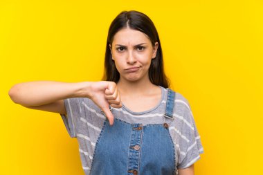 Izole sarı arka plan üzerinde dungarees genç kadın başparmak aşağı işareti gösteren