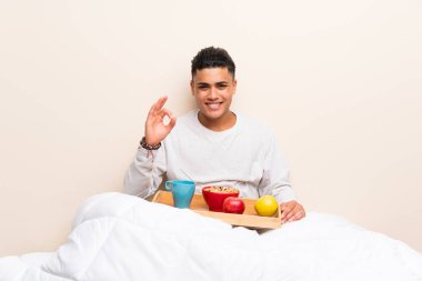 Yatakta kahvaltı olan genç adam parmaklarıyla ok işareti gösteriyor