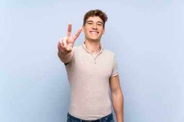 Izole mavi duvar üzerinde Yakışıklı genç adam gülümseyerek ve zafer işareti gösteren