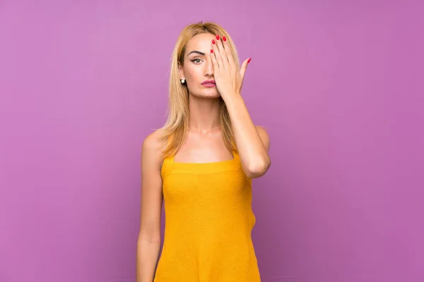 Young blonde woman over purple background covering a eye by hand