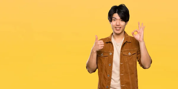 孤立した黄色の背景の上にOkサインと親指アップジェスチャーを示す茶色のジャケットを持つアジアの男性 — ストック写真