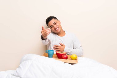 Yatakta kahvaltı eden genç adam davet ediyor