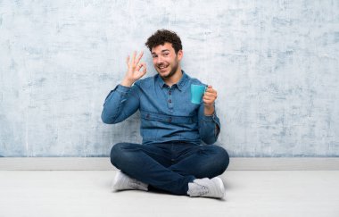 Yerde oturan genç adam sıcak bir fincan kahve tutarak