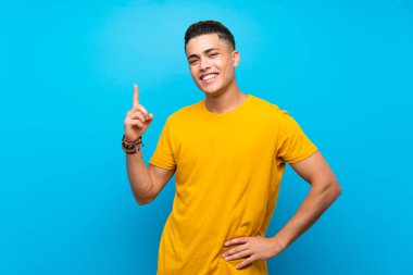 Izole mavi arka plan üzerinde sarı gömlek ile genç adam gösteren ve en iyi işareti bir parmak kaldırma
