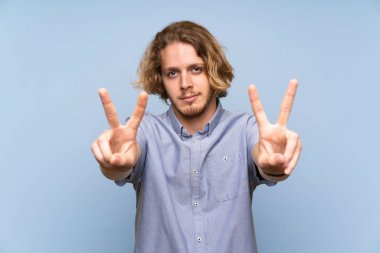 Sarışın adam üzerinde izole mavi duvar gülümseyen ve gösteren zafer işareti