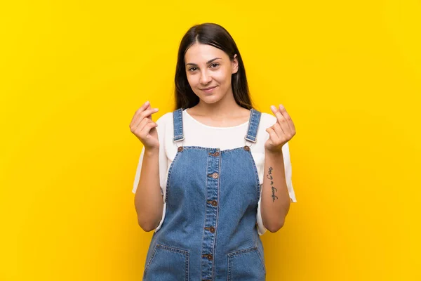 Jovem Mulher Dungarees Sobre Isolado Amarelo Fundo Fazendo Dinheiro Gesto — Fotografia de Stock