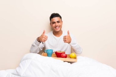 Yatakta kahvaltı yapan genç adam başparmak jesti yapıyor