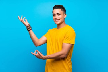 Izole mavi arka plan üzerinde sarı gömlekli genç adam gelmek için davet için yan ellerini uzatarak