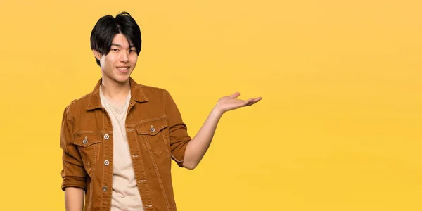 茶色のジャケットを持つアジア人男性が 孤立した黄色の背景に広告を挿入するために手のひらにコピースペースの架空を保持している — ストック写真