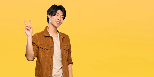 Asian Man Brown Jacket Smiling Showing Victory Sign Isolated Yellow — Stock Photo, Image