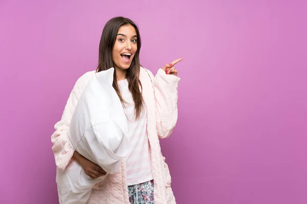 Young woman in pajamas and dressing gown over isolated purple background surprised and pointing finger to the side