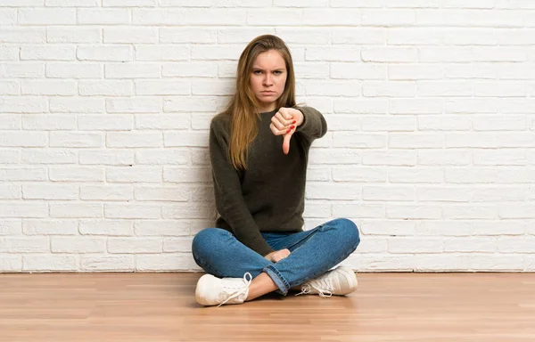 Młoda Kobieta Siedzi Podłodze Pokazując Kciuka Dół Negatywnym Wyrazem — Zdjęcie stockowe