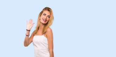 Genç sarışın kadın izole mavi arka plan üzerinde mutlu ifade ile el ile selamlama